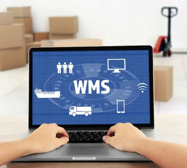 Hands typing on laptop displaying WMS software interface with warehouse boxes background