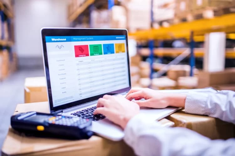 hands typing on a laptop showing a warehouse inventory screen while standing in a warehouse