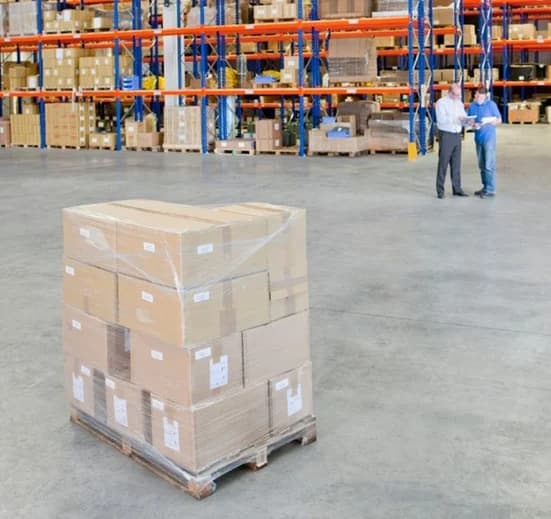 Warehouse workers reviewing inventory next to palletized cardboard boxes on shelving racks