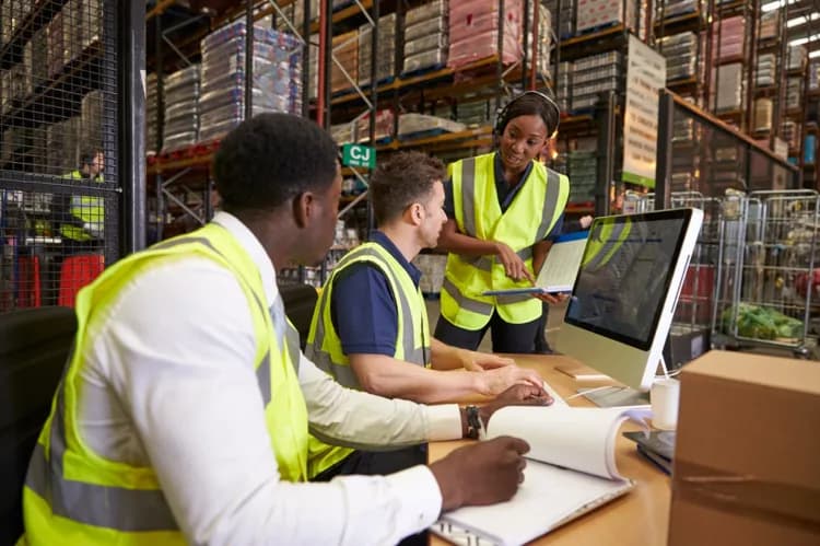Three warehouse workers in safety vests collaborating on shipping processes using computer systems