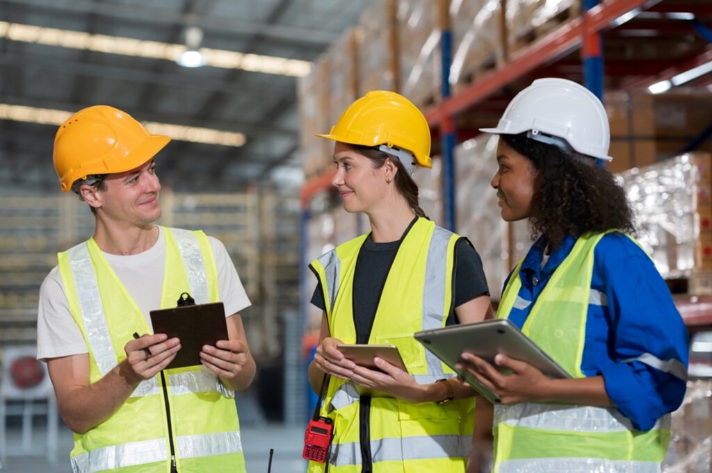 warehouse workers smiling one holding a clipboard and the other two holding a tablet and a laptop